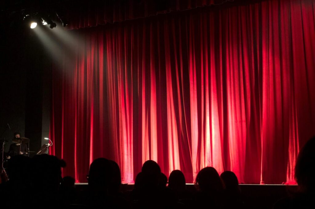 Dimly lit theater stage with red curtains and audience silhouettes under spotlights.
